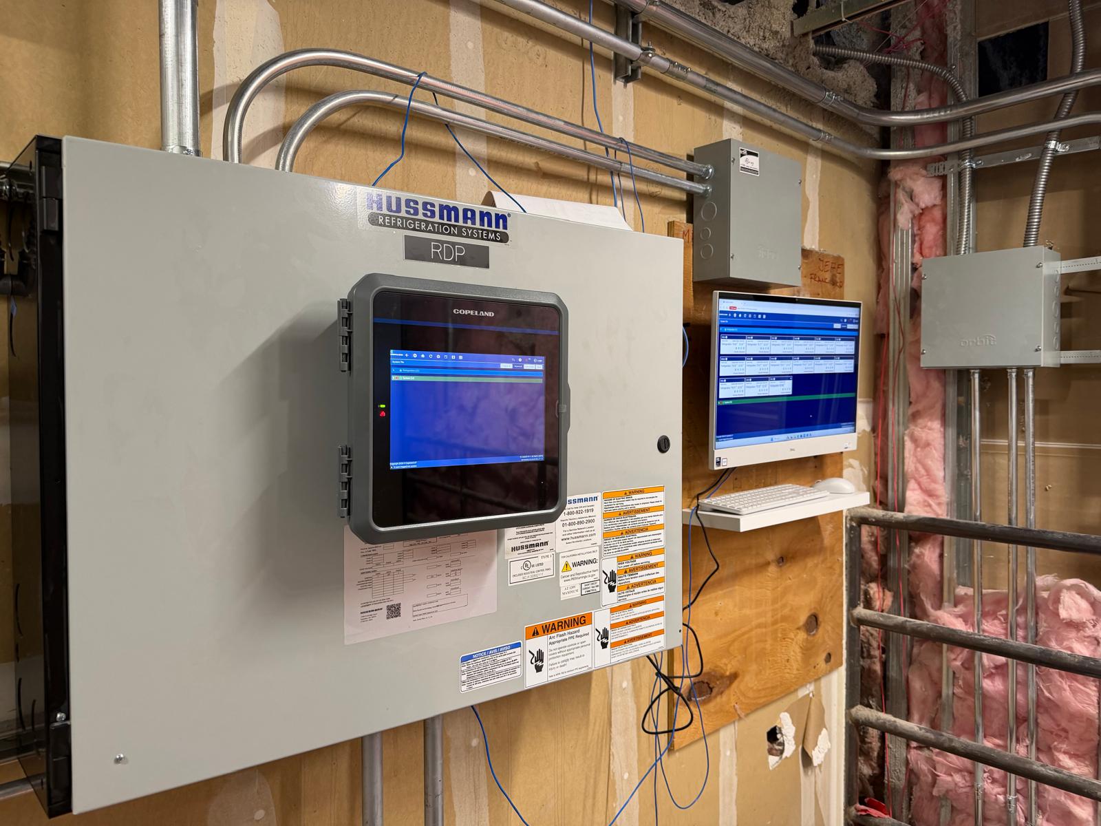 Field installation with Copeland E3 controller mounted on a Hussmann refrigeration panel and adjacent remote monitoring workstation